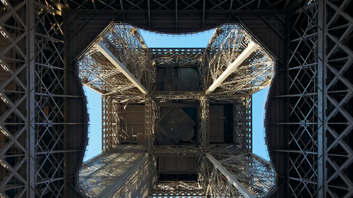 Struktur kisi besi Menara Eiffel difoto dari bawah tepat di tengah keempat kakinya, mendongak ke langit